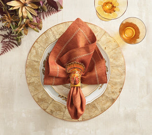 A rust-colored cloth napkin held by a metallic Kim Seybert Turkey Trot Napkin Ring, resting on a white dinner plate and a textured charger for autumn decor.