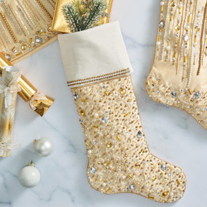 The Kim Seybert Gilded Spray Gold and Crystal Stockings, in shimmering gold and white with sequins and beads, are displayed on marble beside wrapped gifts, ornaments, and a festive holiday accent tree skirt.