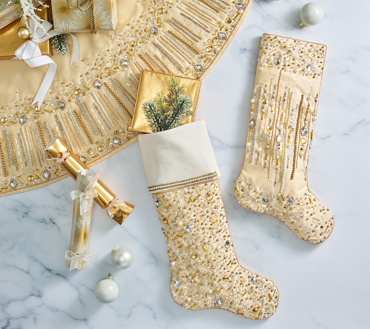 Two gold and white stockings from the Kim Seybert Gilded Sparkle Stocking and Tree Skirt Collection, adorned with sequins and beads, rest on marble alongside metallic ornaments, a wrapped gift, decor accents, and the matching tree skirt.