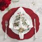 The Christmas Carol Table Linen Collection by Kim Seybert adds elegant embroidered linen detail to a holiday table set with a red placemat, white plate, festive napkin ring, utensils, wine glass, and red roses on light wood.