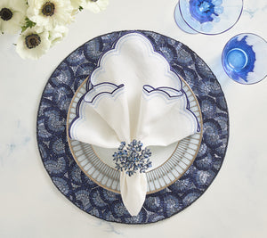 A table setting featuring the Kim Seybert Lune Napkin Set with scalloped edges, a blue and white patterned plate, a blue floral napkin ring, two blue glasses, and white flowers on the side.