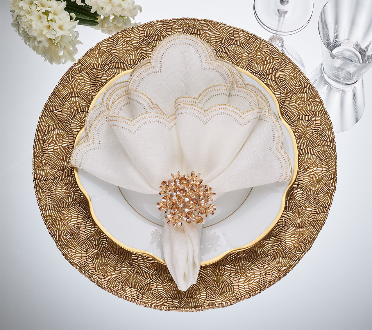 A gold and white table setting highlights a fan-folded Kim Seybert Lune Napkin, secured with a jeweled napkin ring and displayed on a gold charger plate.