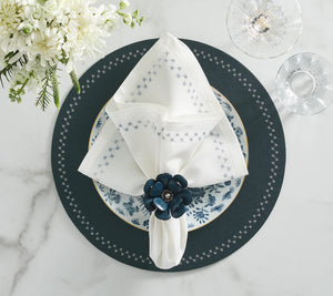 The Kim Seybert Starry Night Napkin Set, featuring white linen napkins and blue floral rings, rests on a blue and white patterned plate atop a dark charger—an elegant table accent with metallic embroidery, glassware, and cutlery on marble.