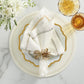 A white napkin with a gold ring sits on a white, gold-rimmed plate atop a Kim Seybert Starry Night Placemat, flanked by two amber glasses and a floral arrangement on a marble surface.