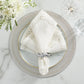 A white embroidered napkin with a silver ring rests on a decorative plate atop the Kim Seybert Starry Night Placemat, next to a glass and white flowers on a marble surface.