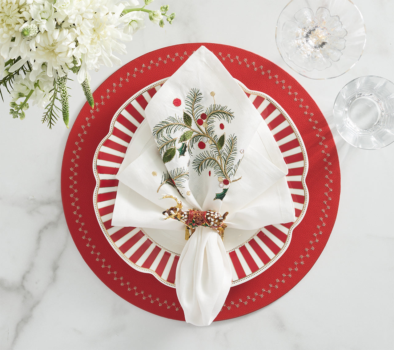 Create a festive holiday table with the Kim Seybert Christmas Carol Table Linen Collection, featuring embroidered napkins in decorative rings, paired with red and white striped plates, clear glassware, and white flowers on marble.
