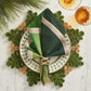 A table setting with a white plate with green leaf pattern, a green napkin and gold ring, Kim Seybert’s Verve Placemat in Green & Natural, pine needles, and two glasses filled with amber liquid.