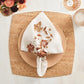 A folded white napkin with embroidered autumn leaves is held by a ring and placed on a patterned plate atop the Kim Seybert Whistler Placemat, adding rustic charm to your table. Glassware and florals complete the look.