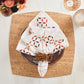 The Kim Seybert Malta Napkin Set, featuring a geometric pattern and beaded napkin ring, is styled on a gold-trimmed brown plate atop a square cork placemat, evoking the refined elegance of cutwork traditions.