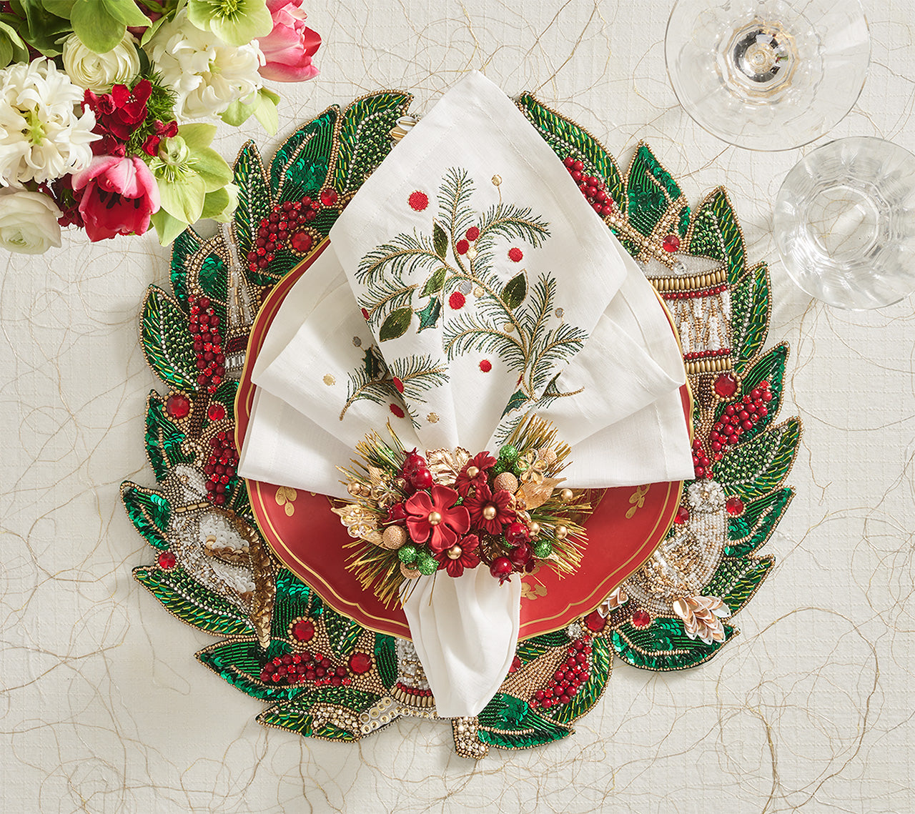 A festive holiday table set with a red plate, embroidered linen napkin, beaded leaf placemat, floral napkin ring, and glassware atop the Kim Seybert Christmas Carol Table Linen Collection.