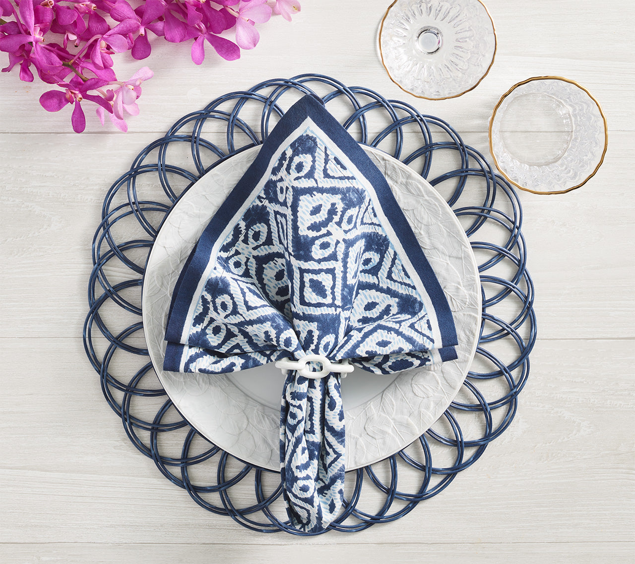 A white plate with a Kim Seybert Alfresco Napkin Set—cotton napkins featuring subtle block printing—rests on a decorative blue placemat, surrounded by glassware and pink flowers on a light tabletop.