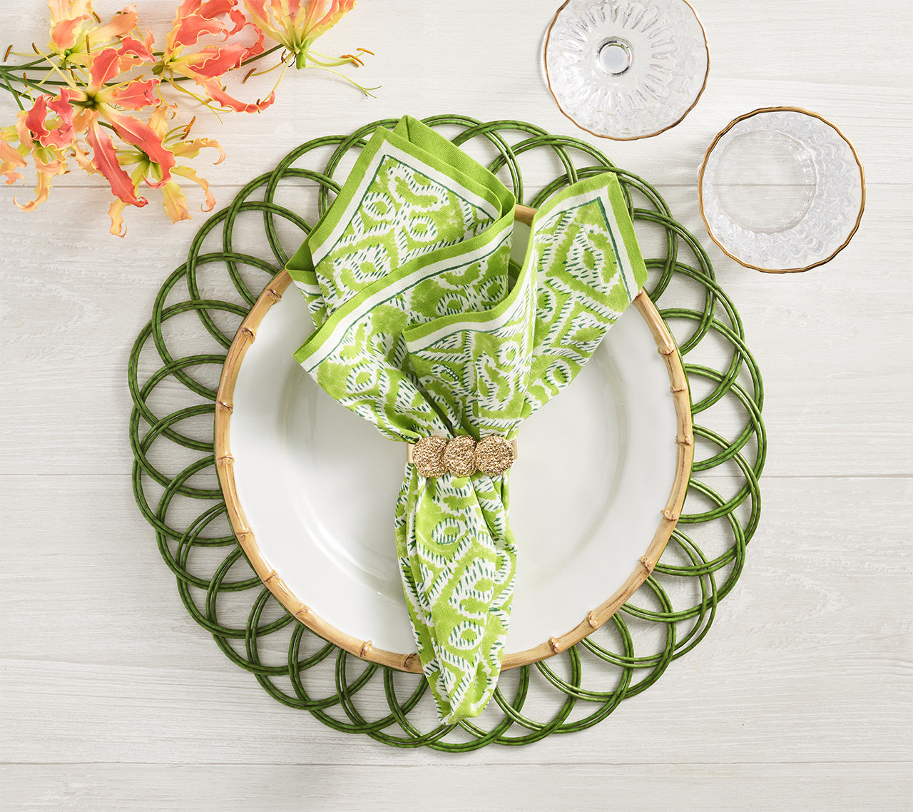 A white plate with a Kim Seybert Alfresco Napkin—cotton with a green and white block print—rests on a green woven placemat, paired with gold-rimmed glassware and orange flowers.