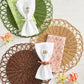 Three handwoven Alfresco Placemats by Kim Seybert, featuring scalloped edges, are arranged with patterned napkins and embroidered napkin rings on a white surface, accented by orange flowers for decoration.