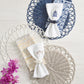 Three Kim Seybert Alfresco Placemats with scalloped edges are displayed on light wood, topped with folded napkins and rings, handwoven faux rattan textures, and purple flowers accenting the lower left corner.
