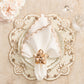 A white plate with a folded napkin in the Kim Seybert Beachcomber Napkin Ring Set rests on a cream, scalloped placemat with shell and bead accents, beside clear glasses and blush flowers—ideal for coastal table decor.