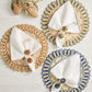 Three round Casbah Placemats by Kim Seybert, including a vibrant green corn husk design, each hold a folded white napkin with a decorative ring and are elegantly arranged on a light wooden surface.