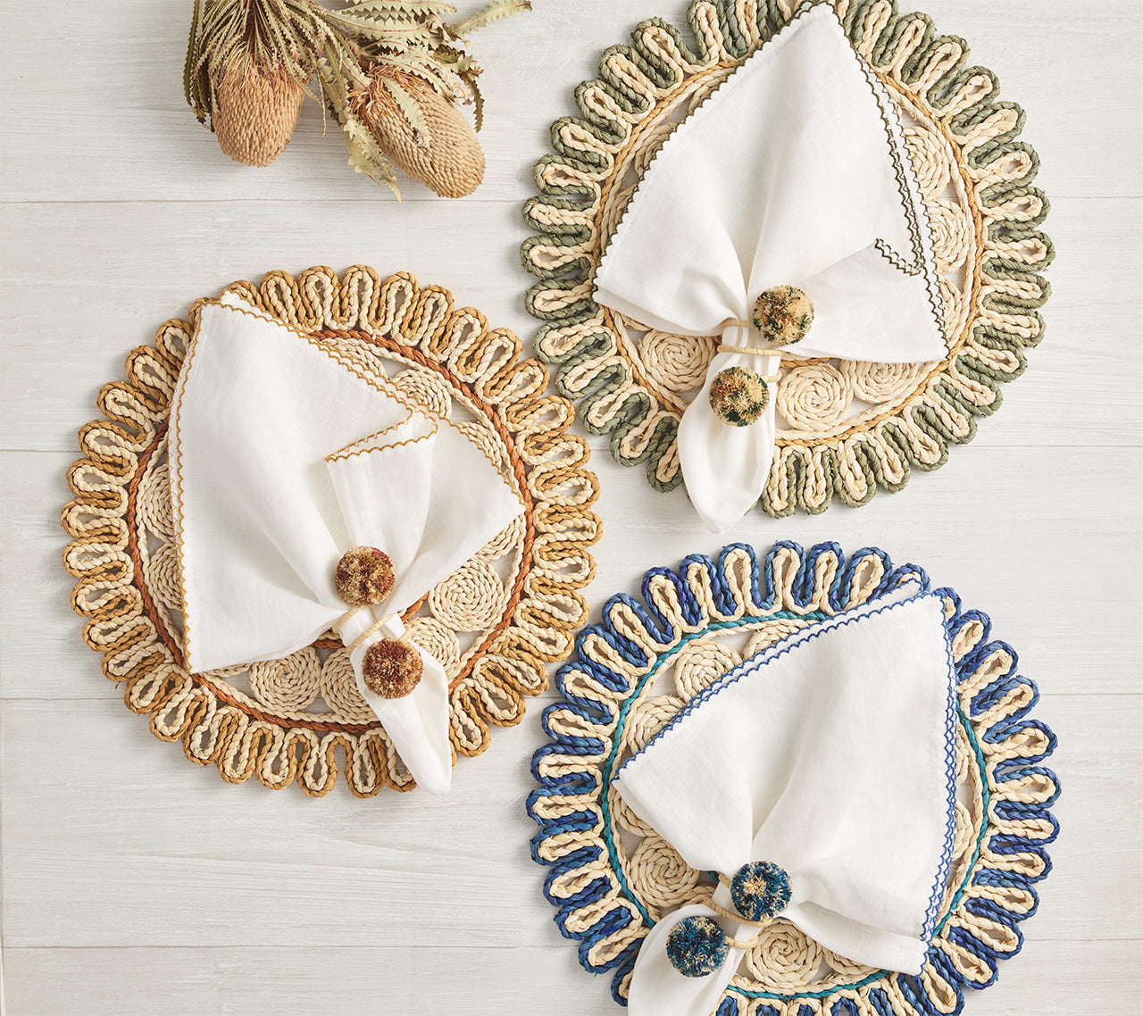 Three round Casbah Placemats by Kim Seybert, including a vibrant green corn husk design, each hold a folded white napkin with a decorative ring and are elegantly arranged on a light wooden surface.