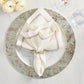 A folded embroidered napkin with a napkin ring rests on a white plate with a silver rim, set atop the Kim Seybert Chalet Placemat, next to a wine glass and white flowers for a nature-inspired table setting.
