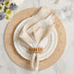 A beige napkin with a Kim Seybert Spoke Napkin Ring rests on layered white plates with dotted edges atop a textured placemat; two empty glasses and a floral arrangement complete the elegant table decor.