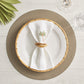 A white plate with a bamboo-style rim sits on a textured placemat. In the center, a white napkin is folded creatively and secured with the Kim Seybert Natural Bamboo Trinity Napkin Ring. Glassware accompanies the setting.