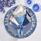 A table setting with blue and white plates, a floral napkin ring, cobalt blue beads, a Kim Seybert Lagoon Placemat, a blue glass, and purple flowers arranged on a marble surface.