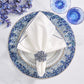 The Lagoon Placemat by Kim Seybert anchors a white napkin with a cobalt blue beaded ring on a patterned plate, alongside blue flowers and glassware—creating an ocean-inspired tablescape.