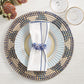 A table setting on light wood features a woven placemat, striped plate, two clear glasses, folded white napkin with blue trim, and a Kim Seybert Pop Rocks Napkin Ring for added sparkle.