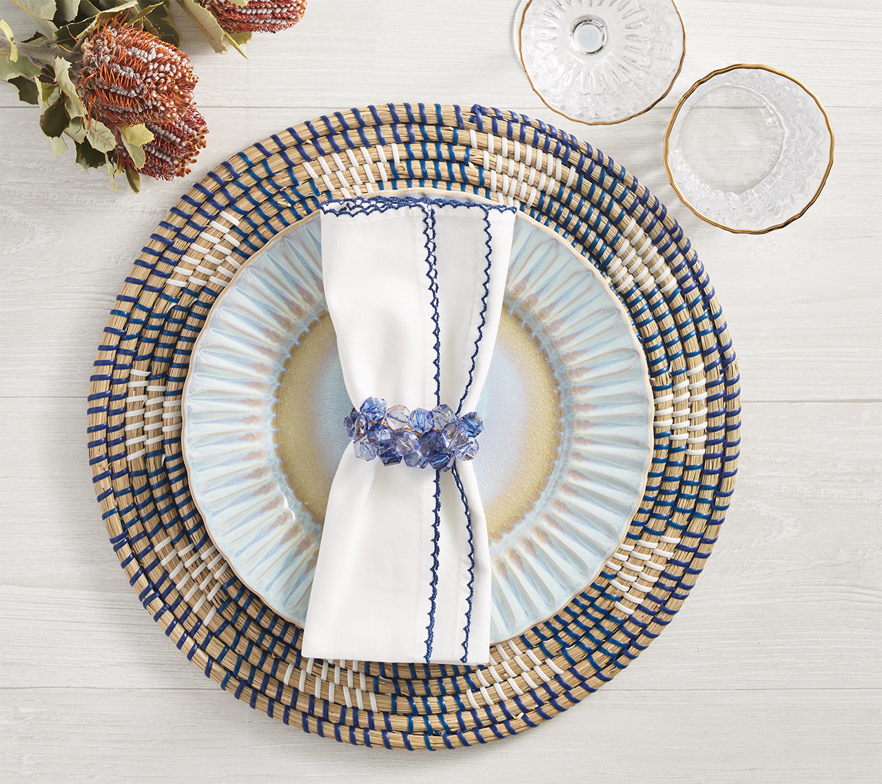 A table setting on light wood features a woven placemat, striped plate, two clear glasses, folded white napkin with blue trim, and a Kim Seybert Pop Rocks Napkin Ring for added sparkle.