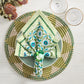 A neatly arranged place setting features a woven placemat, light green plate, Kim Seybert Majorelle Napkin Set with Indian block printing, and two clear glasses on a light wooden table.