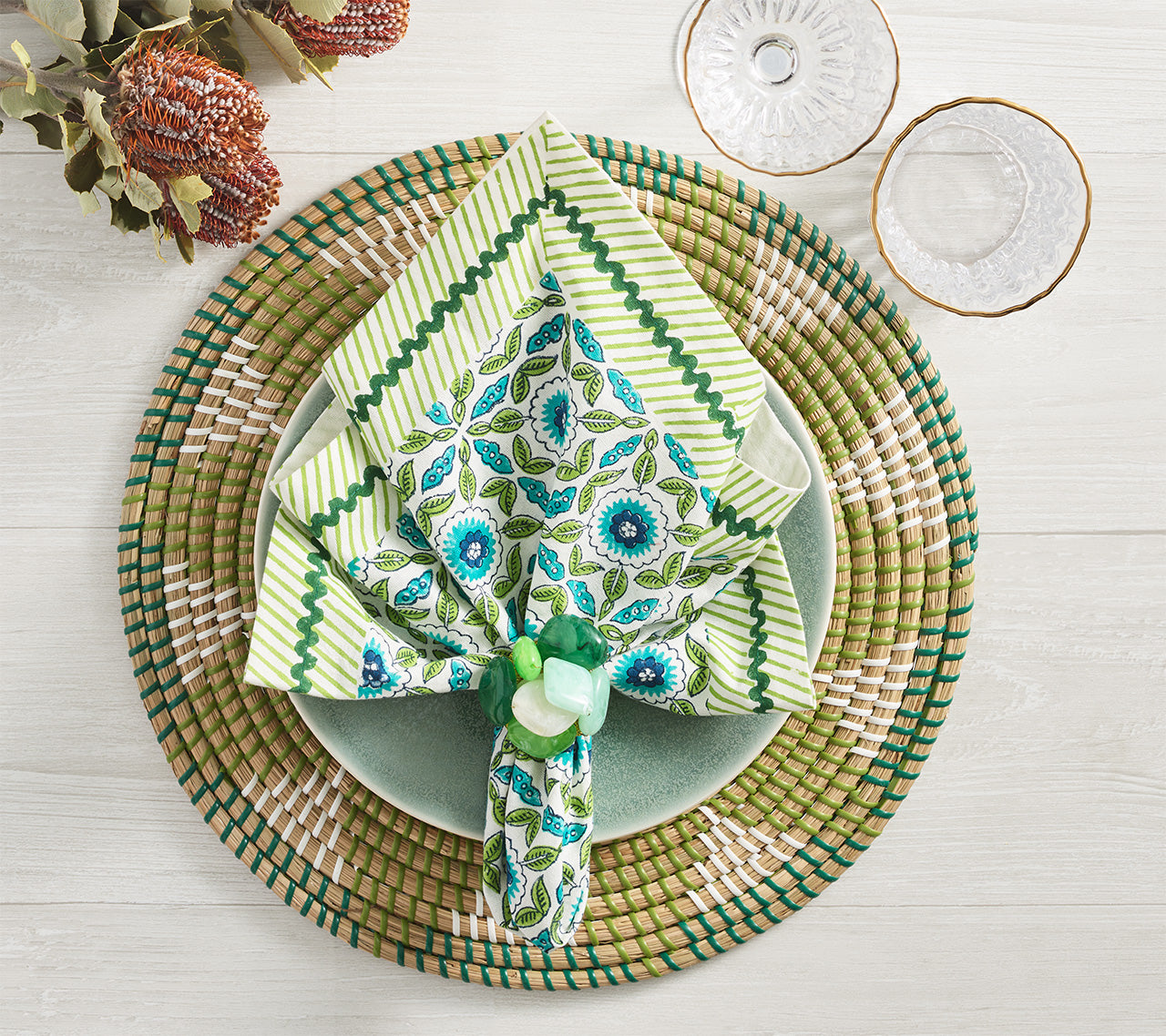 A neatly arranged place setting features a woven placemat, light green plate, Kim Seybert Majorelle Napkin Set with Indian block printing, and two clear glasses on a light wooden table.