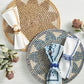 Two Kim Seybert Majorelle Placemats, one tan and one blue, each with a folded napkin and napkin ring, are displayed on a light wooden surface with dried flowers, highlighting their subtle floral motifs.
