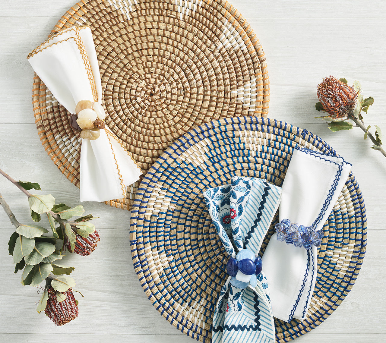 Two Kim Seybert Majorelle Placemats, one tan and one blue, each with a folded napkin and napkin ring, are displayed on a light wooden surface with dried flowers, highlighting their subtle floral motifs.