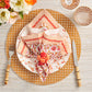 A table setting featuring a patterned plate, Kim Seybert Majorelle Napkin Set in orange and white, a faux fruit napkin ring, bamboo-handled cutlery, stemmed glassware, and a floral centerpiece.