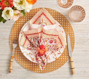 A table setting featuring a patterned plate, Kim Seybert Majorelle Napkin Set in orange and white, a faux fruit napkin ring, bamboo-handled cutlery, stemmed glassware, and a floral centerpiece.