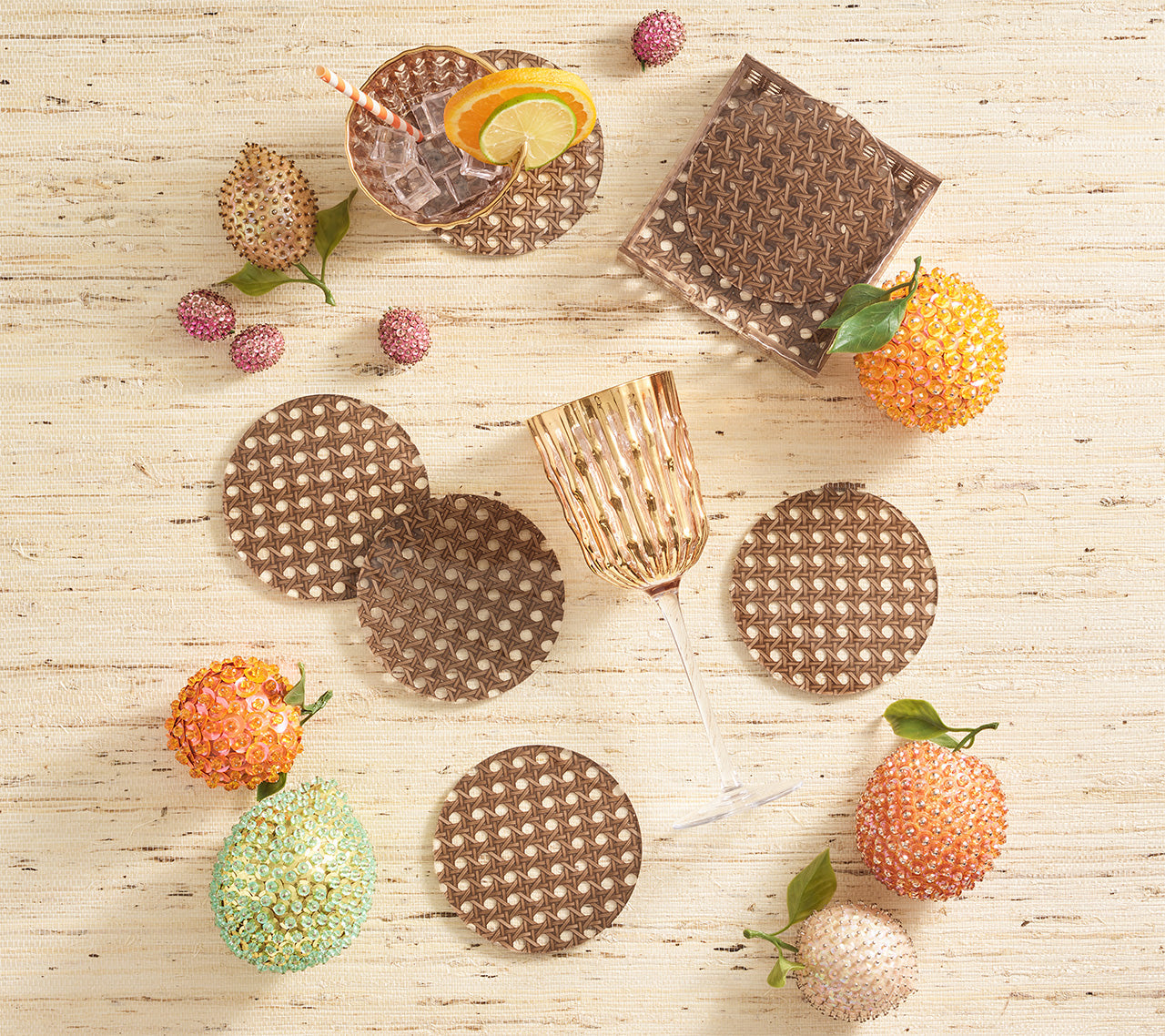 A glass and the Kim Seybert Reed Brown Coaster Set (six faux-wicker coasters) rest on a textured surface, surrounded by colorful beaded fruits and a citrus-garnished drink with a straw.