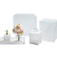 The Kim Seybert White Reed Bath Collection features white perforated accessories with a subtle reed pattern—includes a tray, soap dispenser, tissue box, waste bin, and a vase with assorted flowers.