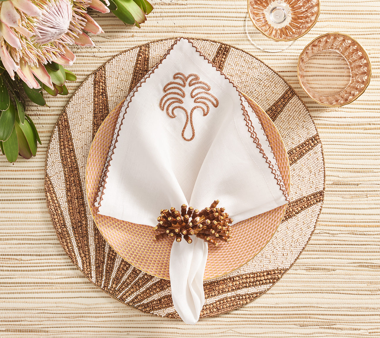 A table setting featuring a white embroidered napkin in a gold ring on a patterned plate, atop the Kim Seybert Royal Palm Placemat, with glassware and flowers on a beige textured tablecloth.