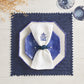 A blue octagonal plate rests on a white plate with a white napkin and blue napkin ring, set atop the Kim Seybert Vista Reversible Placemat. Two blue glasses are placed nearby.