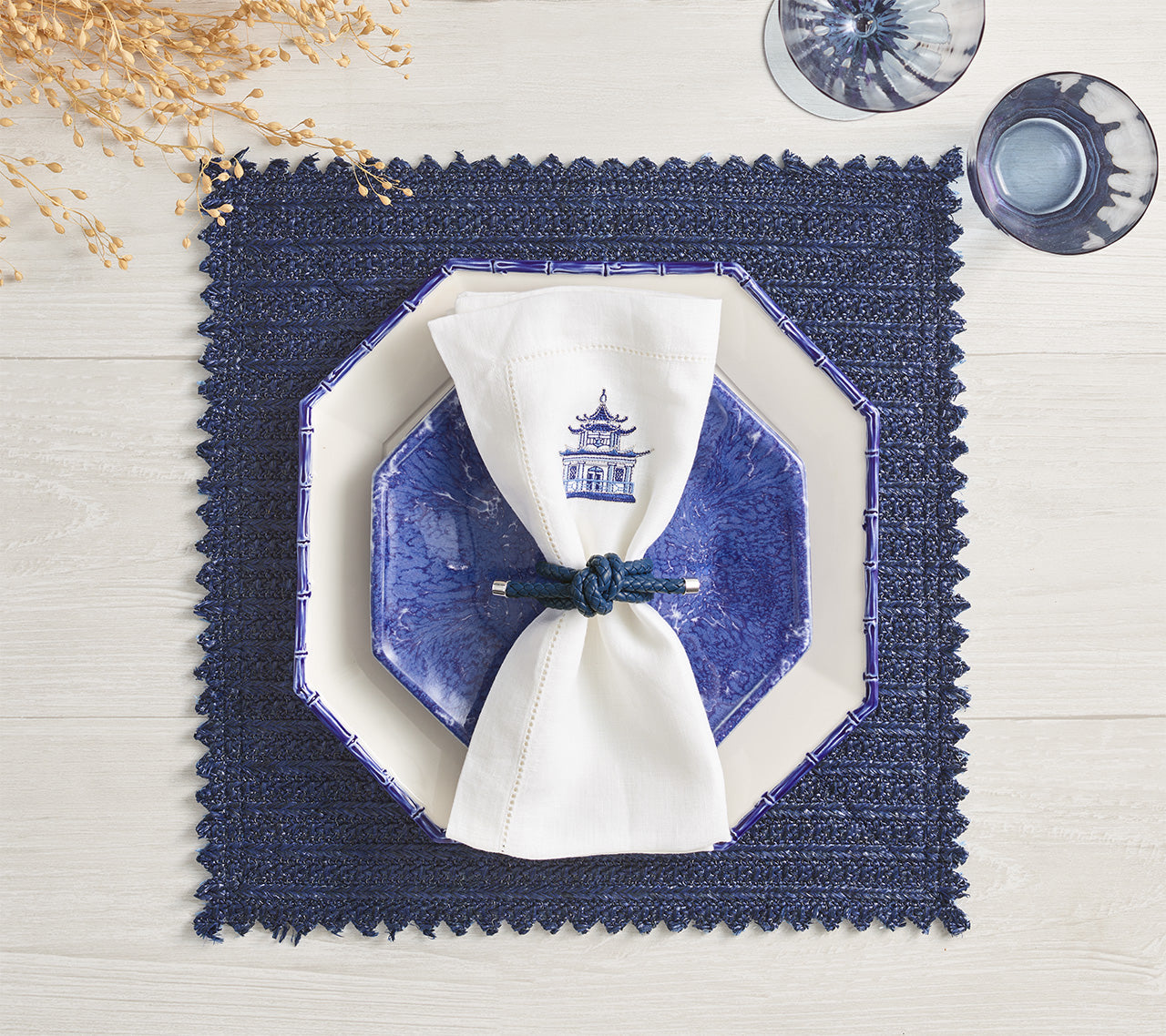 A blue octagonal plate rests on a white plate with a white napkin and blue napkin ring, set atop the Kim Seybert Vista Reversible Placemat. Two blue glasses are placed nearby.
