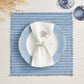 The Kim Seybert Vista Reversible Placemat in blue basketweave is styled with a white plate, blue-edged plate, knotted white napkin, two glasses, and dried stems on a white wooden table.