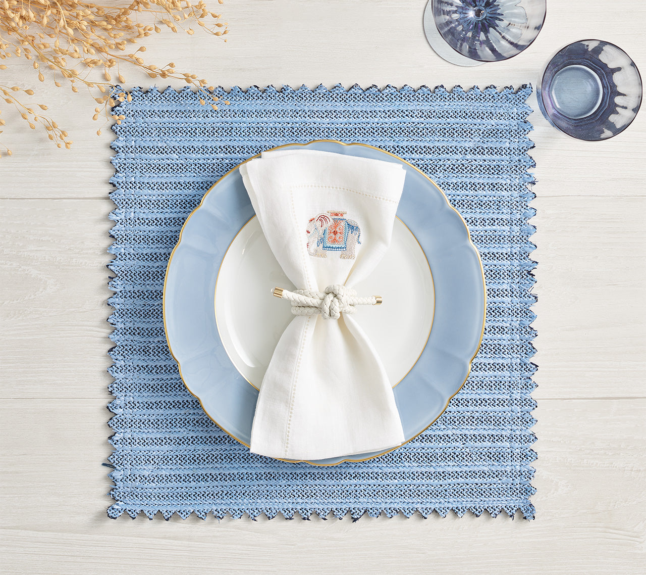 The Kim Seybert Vista Reversible Placemat in blue basketweave is styled with a white plate, blue-edged plate, knotted white napkin, two glasses, and dried stems on a white wooden table.