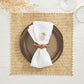 A brown ceramic plate rests on a Kim Seybert Vista Reversible Placemat with classic basketweave, paired with a white napkin featuring pineapple embroidery, a brown napkin ring, and partially visible glassware and dried foliage.
