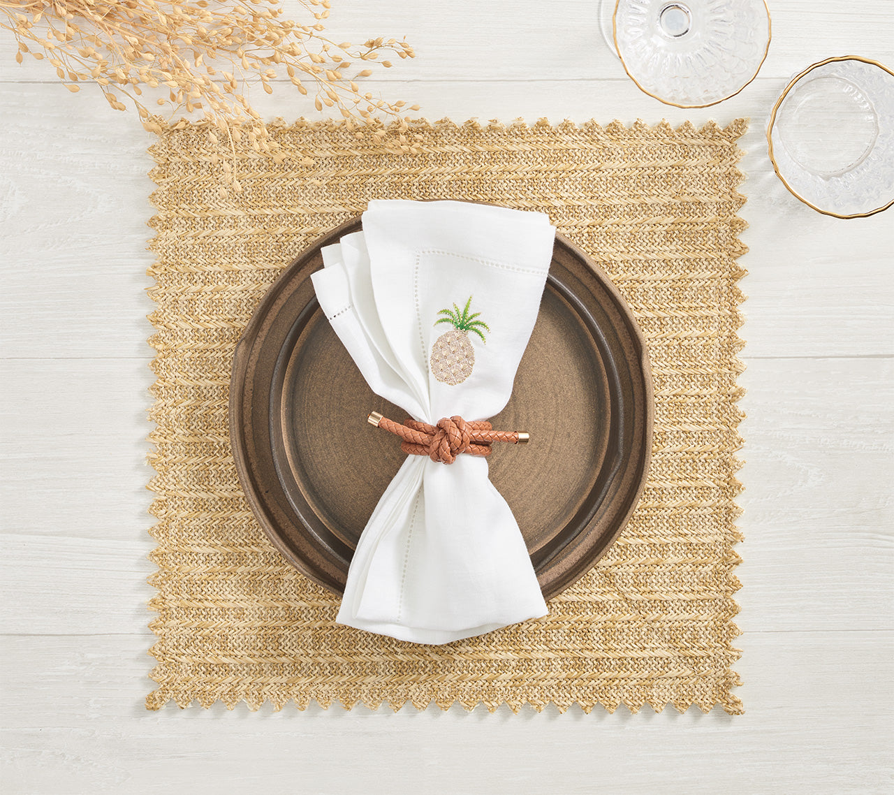 A brown ceramic plate rests on a Kim Seybert Vista Reversible Placemat with classic basketweave, paired with a white napkin featuring pineapple embroidery, a brown napkin ring, and partially visible glassware and dried foliage.