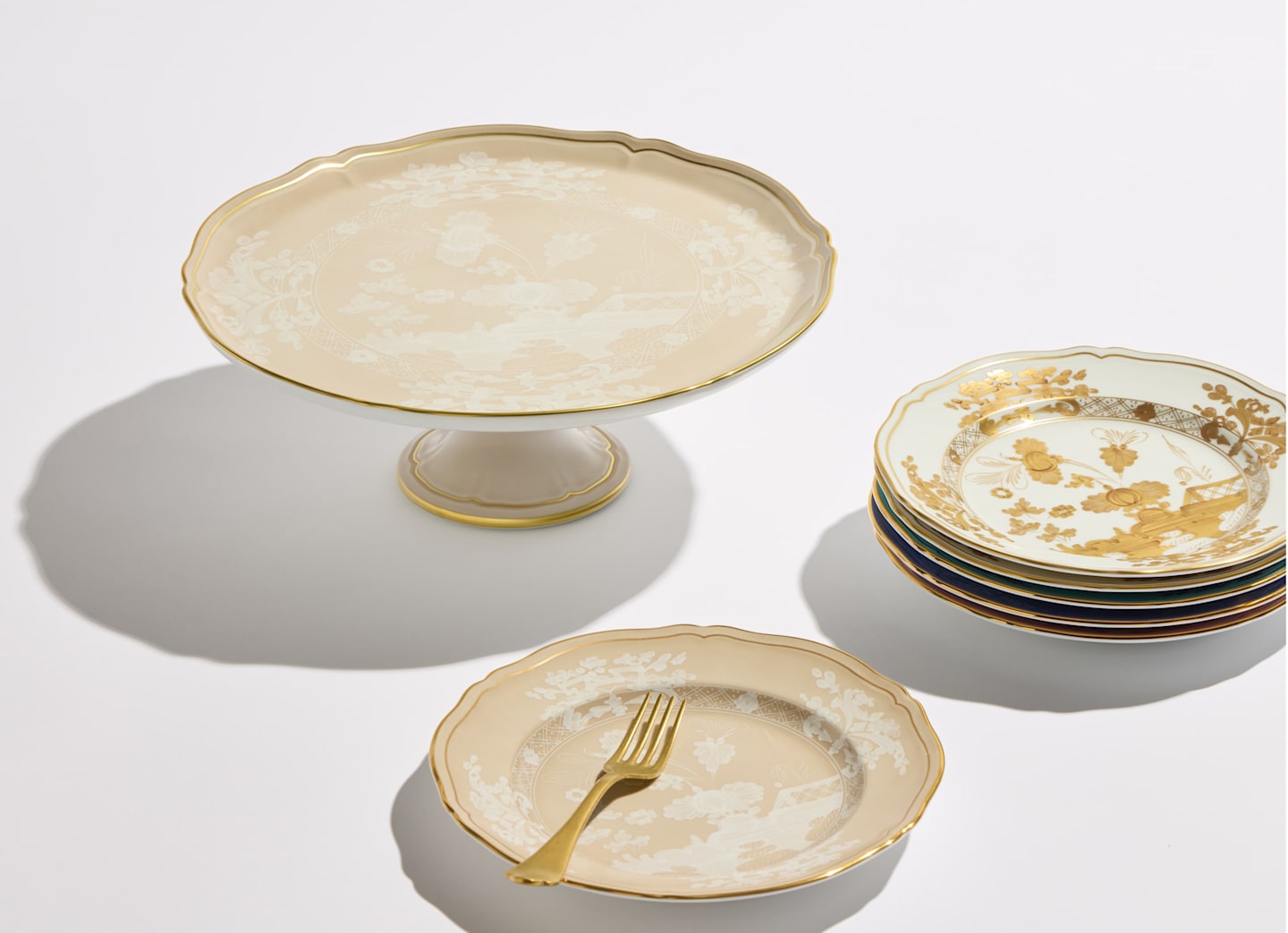 A Ginori 1735 Oriente Italiano Meringa cake stand, stacked dessert plates with gold accents, and a matching plate with a gold fork are artfully arranged on a white surface.