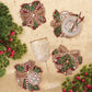 Four Kim Seybert Candy Cane Bow Coasters shaped like bows and decorated with candy canes, holly, and berries, arranged on a gold tablecloth with a wine glass and a small bowl.