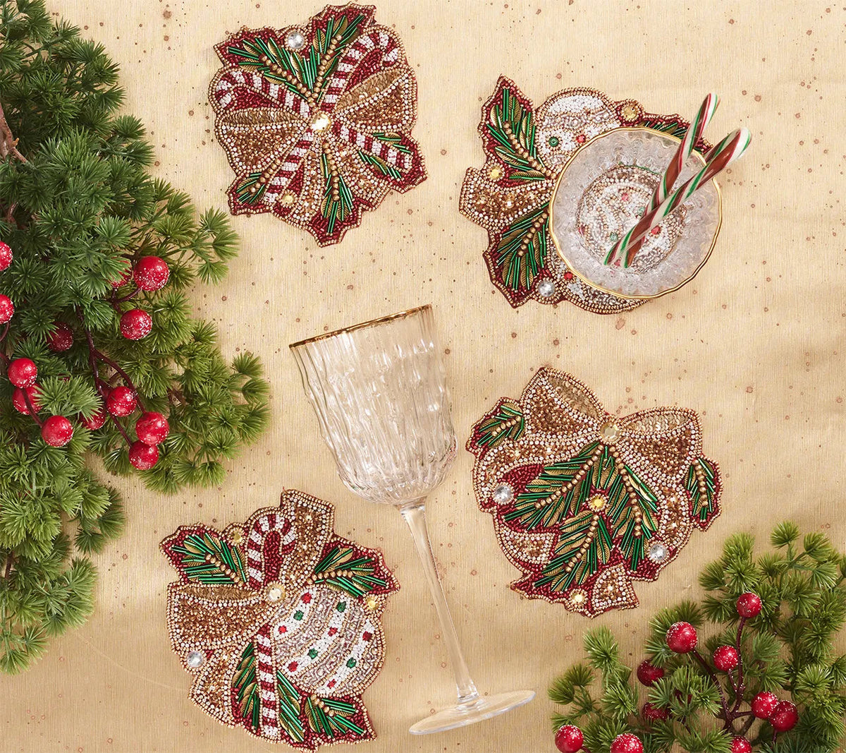 Four Kim Seybert Candy Cane Bow Coasters shaped like bows and decorated with candy canes, holly, and berries, arranged on a gold tablecloth with a wine glass and a small bowl.