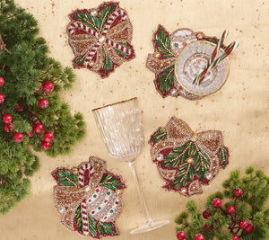 Four Kim Seybert Candy Cane Bow Coasters shaped like bows and decorated with candy canes, holly, and berries, arranged on a gold tablecloth with a wine glass and a small bowl.