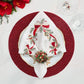 Kim Seybert Tinsel Bow napkin with white embroidery, Christmas ribbons, and ornaments, held by a festive beaded napkin ring on a white plate and red beaded charger.