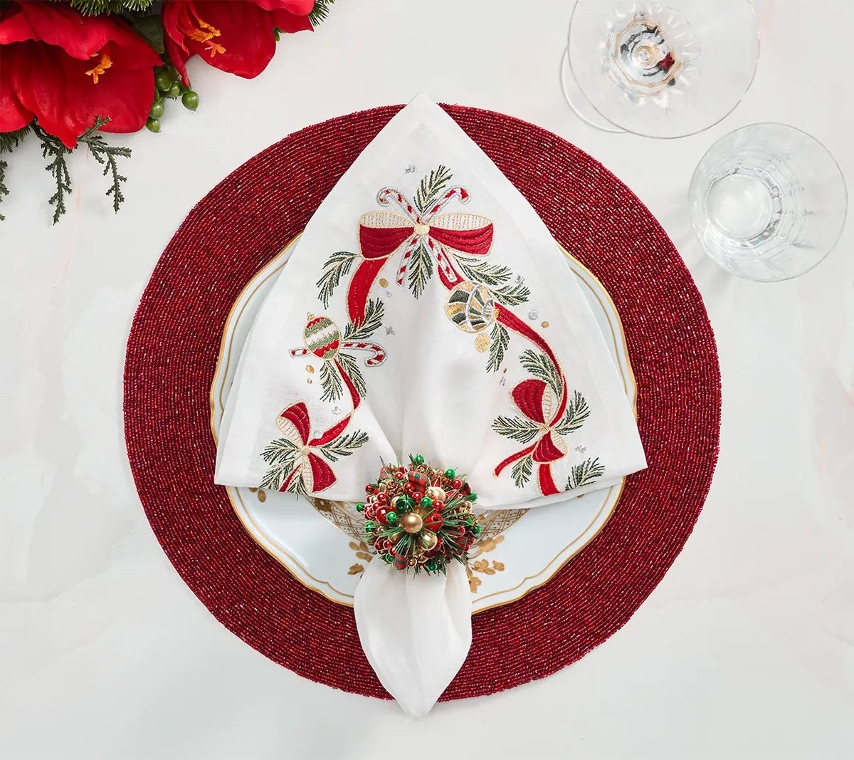 Kim Seybert Tinsel Bow napkin with white embroidery, Christmas ribbons, and ornaments, held by a festive beaded napkin ring on a white plate and red beaded charger.