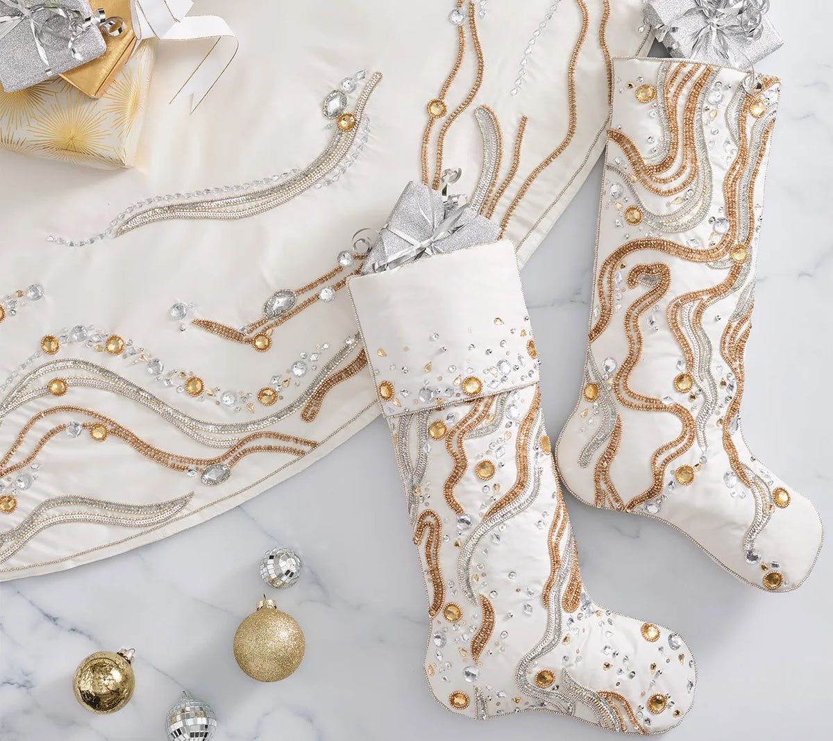 A white Kim Seybert Gilt Wave Tree Skirt and two stockings decorated with gold and silver beadwork, sequins, and crystals, arranged with wrapped holiday décor and ornaments.